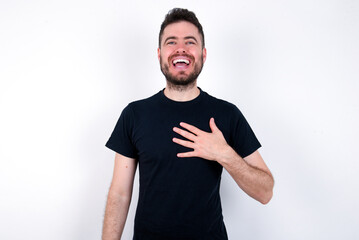 young caucasian bearded man wearing black t-shirt standing over white wall smiles toothily cannot believe eyes expresses good emotions and surprisement