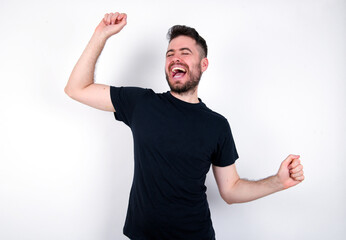 Photo of upbeat young caucasian bearded man wearing black t-shirt standing over white wall has fun...