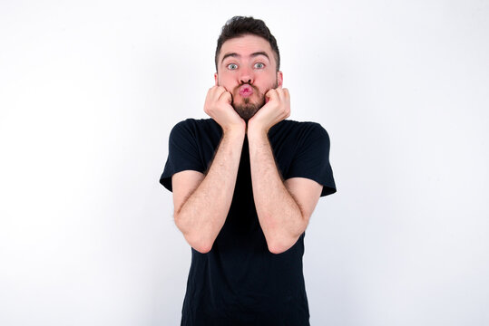 young caucasian bearded man wearing black t-shirt standing over white wall with surprised expression keeps hands under chin keeps lips folded makes funny grimace