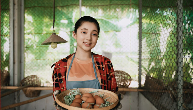 Portrait of happy Asian teenager girl farmer standing holding eggs basket and looking at camera in chicken coop at organic farm.