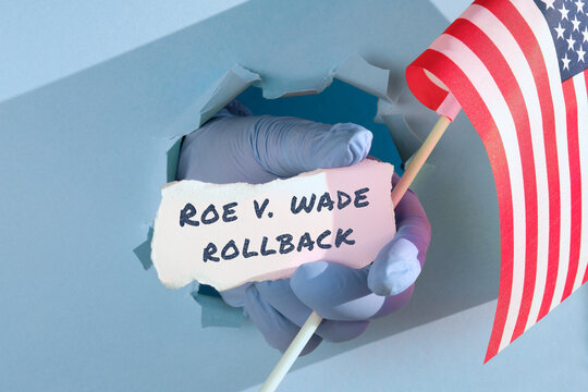 Text Roe vs Wade Rollback on scrap of paper and flag of United States of America. Medics, doctors hand in glove holding USA american flag and scrap of paper text through paper hole.