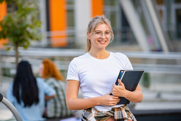 Portrait of a Beautiful smiling blonde hair student girl with glasses  holding books in front of a university building