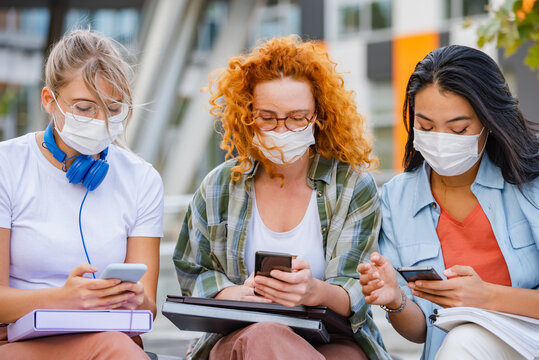 A Diverse Group Of Female College Students Are Sitting In Front Of A University Building Wearing A Protective Face Masks, And Using Smartphones. 