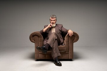Full length shot of pensive mature businessman with beard wearing elegant suit looking at camera, sitting in armchair while posing isolated over light gray background - Powered by Adobe