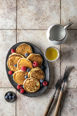 Pancakes on oval plate with fresh berries, honey and rustic cutlery on pink tile background, top view