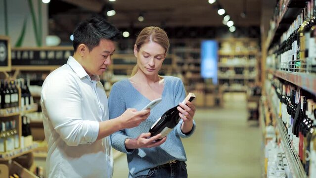 Asian couple chooses wine in a supermarket or grocery store in the alcohol department using a mobile phone smartphone app. husband and wife, man and woman scan the label through use application