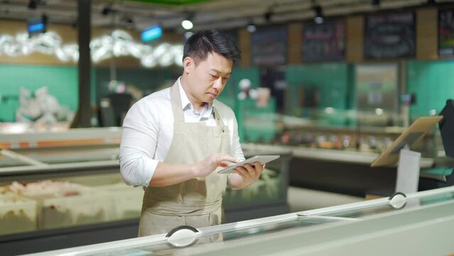 Asian grocery store manager salesman in apron using digital tablet counting goods in supermarket. Male worker employee doing inspection of products regular inventory to control goods, online order