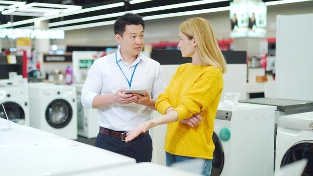 Asian male consultant Salesman Gives Professional advises female home appliance buyer in store. Shop assistant explaining washing machine to customer. showing modern electric 