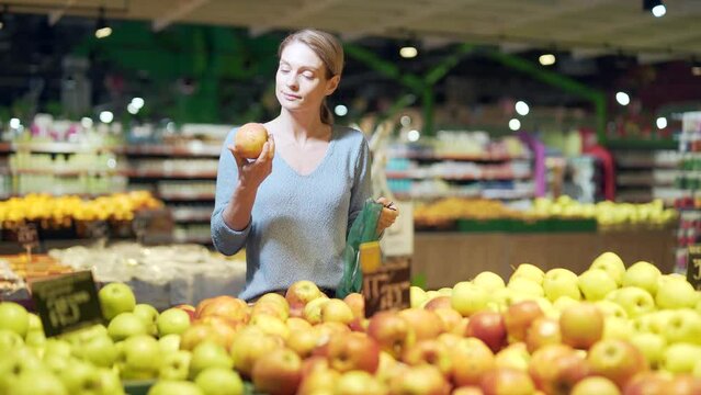 Young Woman Chooses And Picks In Eco Bag Vegetables Or Fruits In Supermarket. Female Customer Standing A Grocery Store Near The Counter Buys And Throws In A Reusable Package In Market Make A Purchase