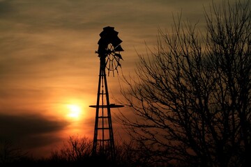 Kansas Sunset with a Windmill silhouette with trees and Sun with clouds north of Hutchinson Kansas USA out in the country.
