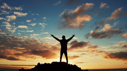 Man stands on a rock against the ocean during summer sunset. Human with raised hands looks to sun...