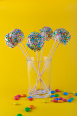 Round homemade sweets with colorful sprinkles stay on sticks in transparent glass. Perty birthday food. Yellow background, vertical photo. Selective focus