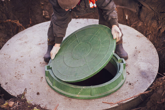 A worker lifts the manhole cover of a sewer well. Construction and maintenance of septic tanks in the village