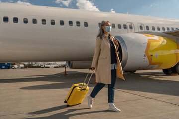 Passenger woman in mask with luggage is outdoors near plane looking away