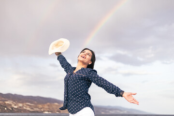 woman smiling with a hat on her hand and with a rainbow on the sky