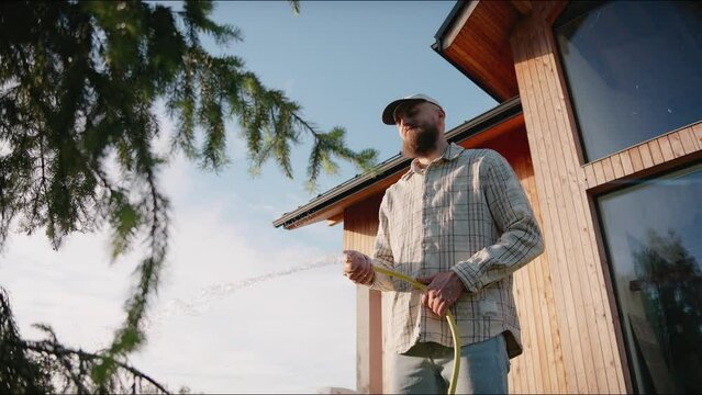Man waters plants and trees with a hose at summer sunset in the backyard. Man takes care of fir tree on a hot summer day in a garden. Watering plants. House yard activities. High quality 4k footage