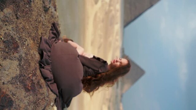 Vertical video. Young redhead girl in brown dress sitting on a stone cliff in Egypt, Cairo - Giza. Pyramids on backround