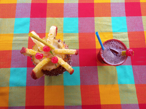 Refreshing Drinks On A Table With A Colorful Tablecloth Is A Piña Colada And A Michelada With A Straw And A Cherry Seen From Above
