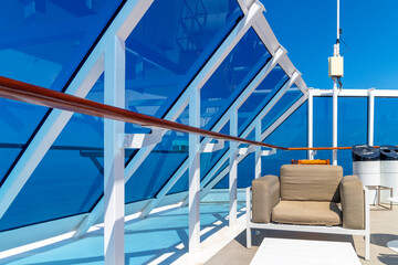 A seating area with glass panels and white railings with a deep blue sky on the upper promenade deck of a cruise ship at sea on a summer day.