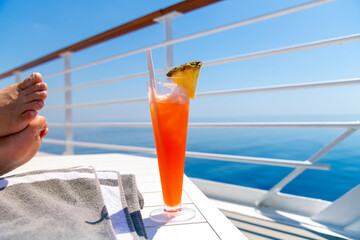 A woman rests her feet on a table on the upper deck of a cruise ship at sea with a colorful drink with pineapple on a hot summer day on the ocean. - obrazy, fototapety, plakaty