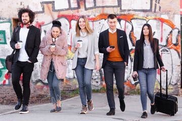 Group of people walking on a street with confidence. Businessmen and businesswomen traveling together. Old wall with abstract unrecognizable graffiti in the background..