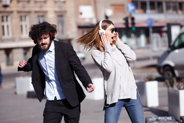 Young business colleagues dancing together with headphones after work. Happy friends jumping and fooling around outdoors