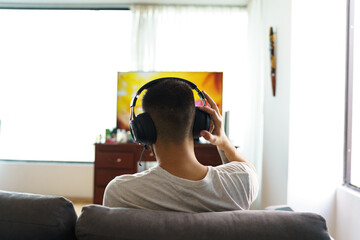 person from the back sitting on a sofa in the living room of a house, using wireless headphones to...