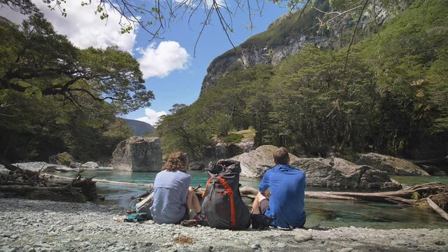 Static, hikers eat lunch on rocky beach overlooking tranquil river, Routeburn Track New Zealand