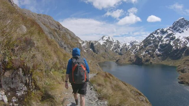 Follow, hiker walks above alpine lake, snow capped mountain landscape, Routeburn Track New Zealand
