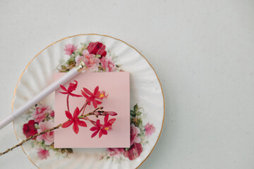 Pink sticky note on floral plate with rose pen and orchid flowers on white background flat lay