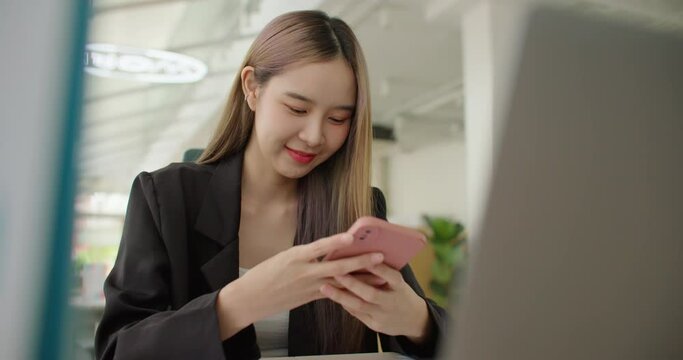 Beautiful smiling asian businesswoman sitting in modern office using smartphone texting, sharing success messages on social media. Attractive happy business freelance woman reading good news on phone.