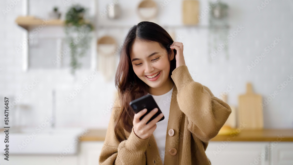 Poster happy young asian woman relaxing at home. asia female standing at counter kitchen and using mobile s