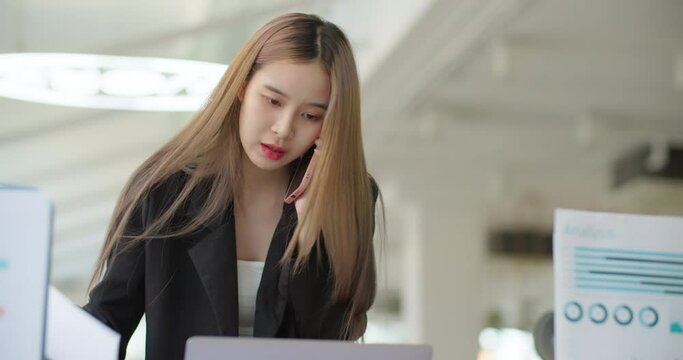 Young busy asian business woman working on project paperwork talking phone, using laptop remotely work in modern office. Female freelancer entrepreneur making distance job telesale call with customer.