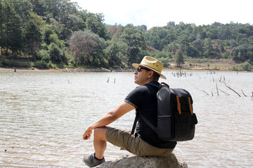 Latin adult man with hat and sunglasses sitting on a stone next to a lake makes the victory sign happy, accomplished, with achievements and goals achieved
