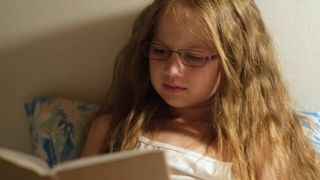 Child girl in glasses reading a book in bed before going to sleep with lamp. Dark bedroom with night light. Children read before bedtime. Childhood, fairy tales and kids dreams