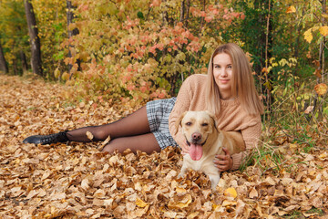 A young woman laying on the dry yellow leaves with her beloved labrador dog in autumn park.