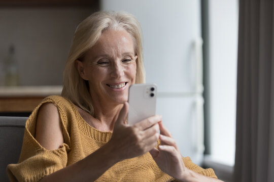 Smiling senior woman spend weekend leisure on internet, relaxing on sofa looks at cell phone screen, watch videos, enjoy in-line content rest at home. Older generation use modern wireless tech concept