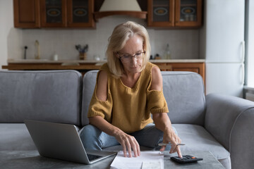 Serious mature 50s woman sit at table calculates household finances, paying bills on laptop use...