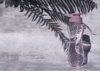 A flat shot of a plastic reusable bottle with an isotonic drink on a gray concrete background with a shadow from palm leaves, hard shadows from the sun, a place to copy.