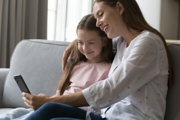 Smiling attractive woman her cute preschool daughter sit on sofa at home, spend free weekend time have fun, play on-line games use modern smartphone. Leisure, young generation and tech overuse concept