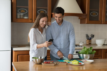 Beautiful couple cooking together vegetable dish use cellphone making food order, delivery at home, enjoy mobile calorie tracking app for comfort life. Modern tech, cookbook recipes on-line concept - Powered by Adobe