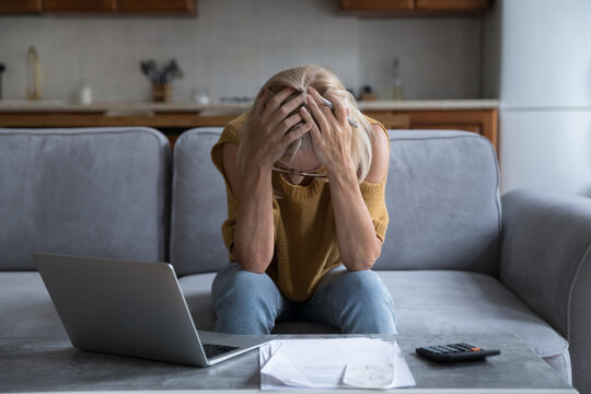 Older Woman Looks Stressed Due Financial Problem Sit On Sofa Near Bills, Calculator, Laptop, Holds Head In Hands, Feels Hopeless. Late Loan Payment, Bankruptcy, Lack Of Money To Pay Utilities Concept