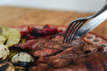 Close shot of cutlery cutting a piece of meat. Traditional Mexican dish with meat. High quality photo. Nopal, sausages and onions. Fork and knife. Wooden table. Enjoying a traditional dish. Delicious