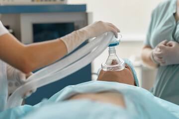 Close up hands of doctor anesthesiologist holding breathing mask on patient face during operation - obrazy, fototapety, plakaty