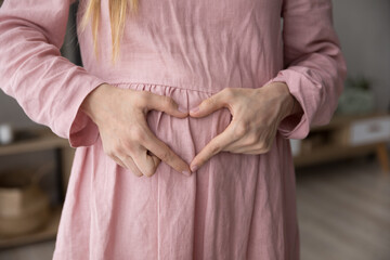 Close up shot unknown pregnant woman in pink dress make heart shape symbol with folded fingers on...