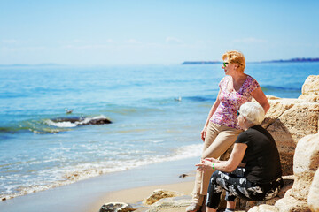 Two elderly women are happy to meet each other, sitting on a rock on the seashore, talking and laughing together. Senior women are happy to communicate