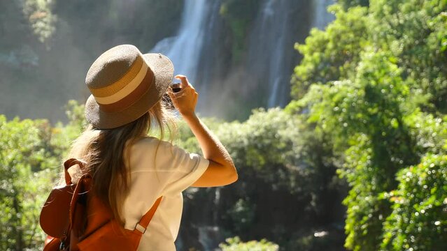Travel woman in Asia journey. Female traveler with photo camera shooting beautiful tropical waterfall. Cinematic video of young tourist girl in wanderlust trip. Discovery famous place.