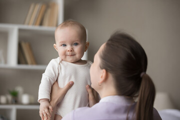 Rear back view young loving mom holding in arms her cute newborn, baby looks at camera. Family enjoy playtime together after daytime nap at home. Parent care, mothers love, infancy, motherhood concept