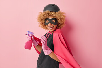 Positive curly female superhero dressed in protective helmet cloak mask and rubber gloves holds spray detergent for cleaning and disinfecting stands against pink background. Domestic chores.