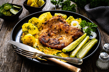 Fried pork chop with bone and egg, boiled potatoes,  cucumber on wooden table
